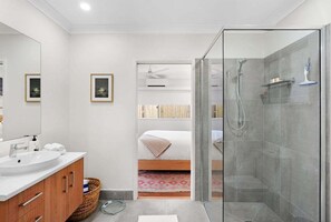 Un baño moderno con cabina de ducha de vidrio, un lavamanos blanco y una cama visible a través de una puerta abierta.