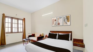 A hotel room with a bed, a chair, a nightstand, a bedside table, a window with curtains, and wall art. - Classic Hotel Phoenix (Valappadi)