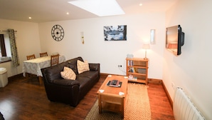 A living room with a brown leather sofa, a wooden coffee table, a dining table with chairs, a clock, a framed picture, a lamp, and a television mounted on the wall. - Milecastle, Irthington , ideal for couples or families exploring Hadrians Wall (Irthington)
