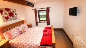 A bedroom with a wooden bed, floral bedding, a window with curtains, a mounted TV, and a radiator. - Milecastle, Irthington , ideal for couples or families exploring Hadrians Wall (Irthington)