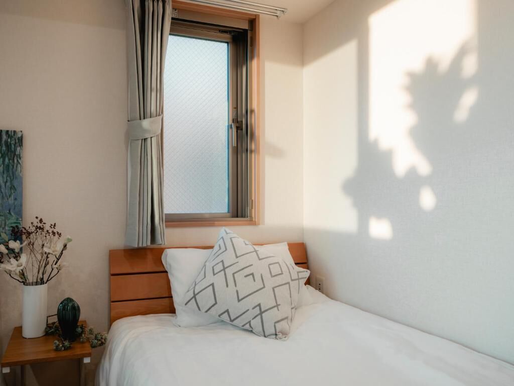 A bedroom with a wooden headboard, a window with curtains, a bedside table with a vase of dried flowers, and a patterned pillow.