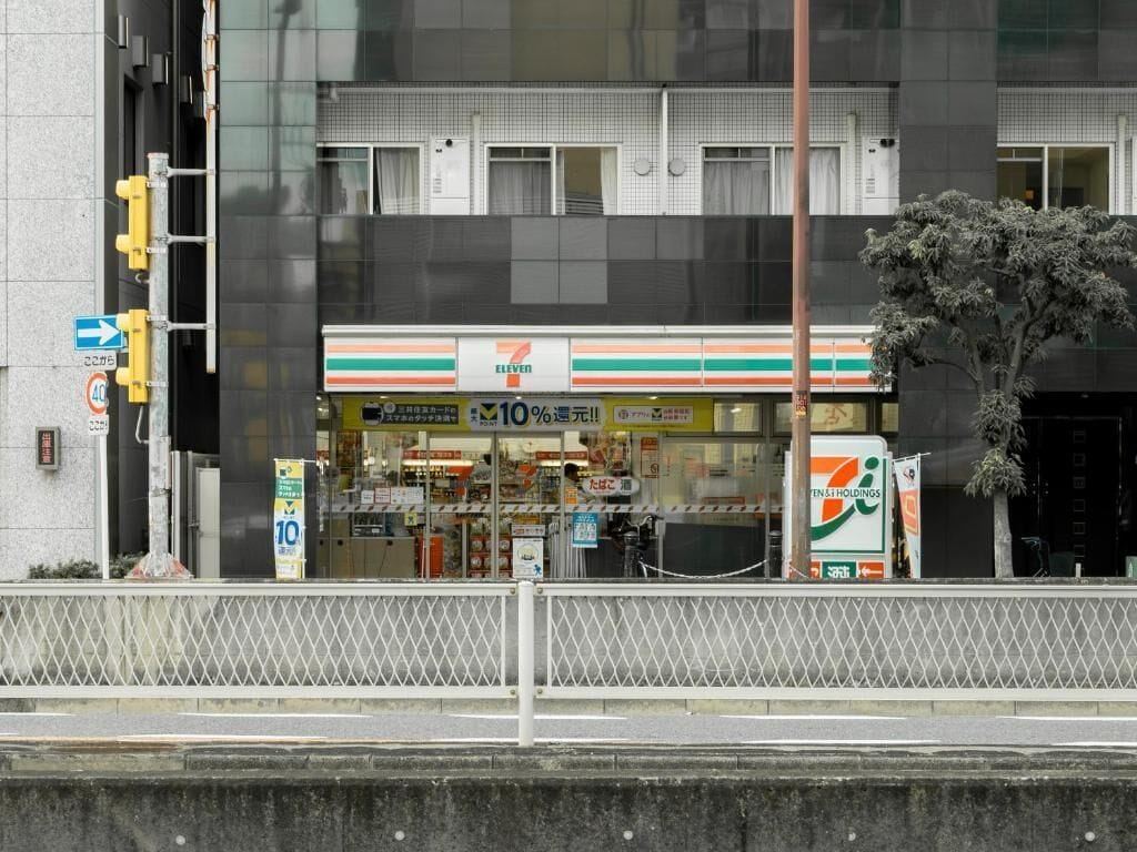 A 7-Eleven convenience store with a 10% off sign, located in front of a modern building with a metal fence in the foreground.