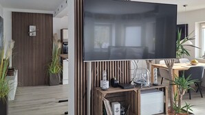 Ein modernes Wohnzimmer mit einem großen Flachbildfernseher, der an einer Wand aus Holzpaneelen befestigt ist. Es gibt einen Essbereich mit Tisch und Stühlen, und verschiedene dekorative Gegenstände stehen auf dem Tisch.
