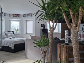 Une chambre moderne avec un lit, un canapé gris et une table d’appoint en bois. On y trouve des fenêtres avec des stores, une bibliothèque et une plante en pot.