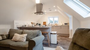 A modern loft apartment with a living area featuring a sofa, a chair, and a small coffee table. A kitchen with a bar counter and a window is visible in the background. - Beautiful 2 Bedroom Apartment in Malvern (Malvern)