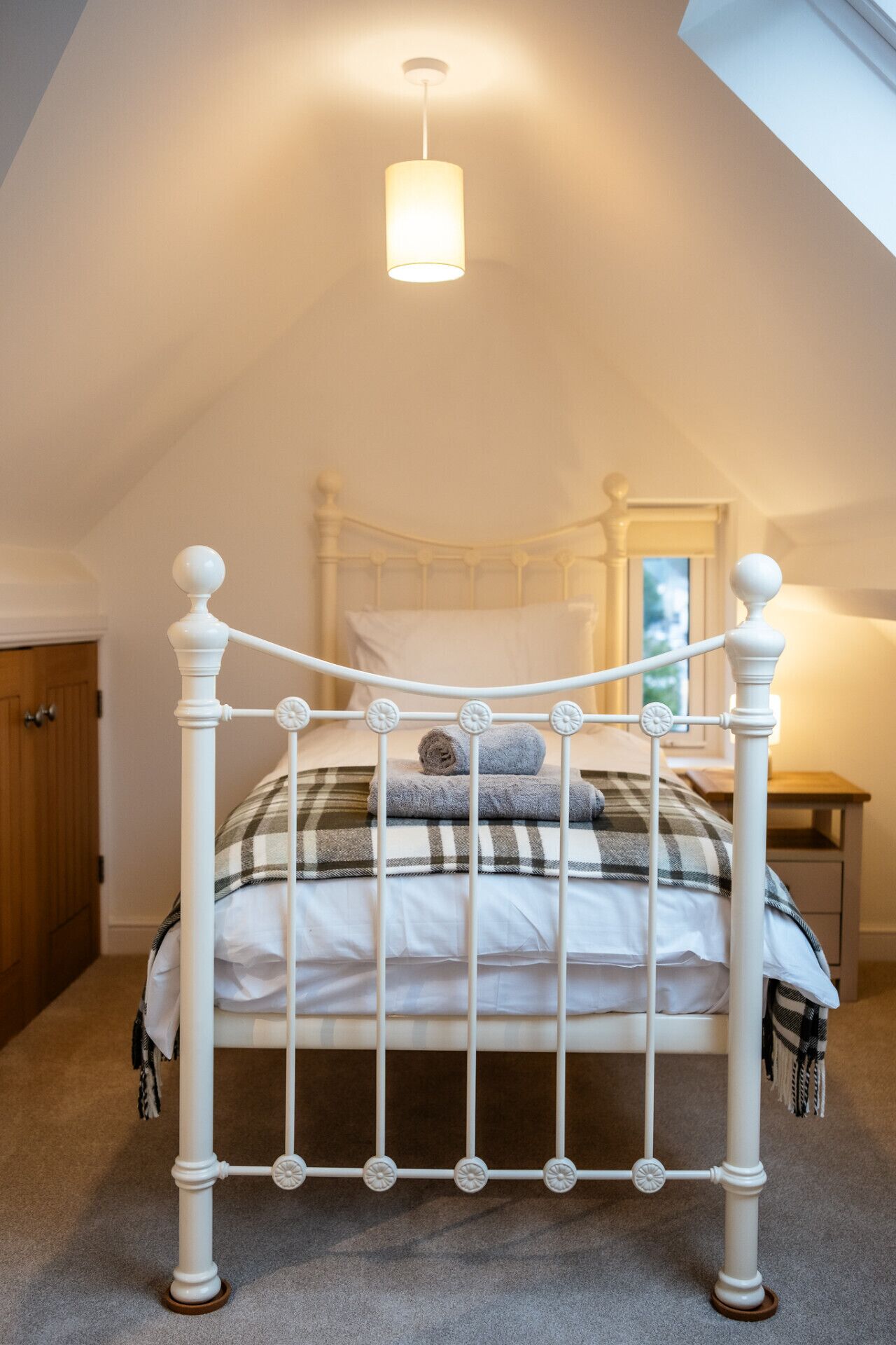 A white metal four-poster bed with a plaid blanket and rolled towels, a bedside table, and a hanging light fixture in a room with a sloped ceiling.