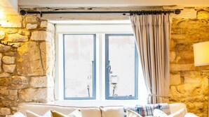 A cozy living room with a stone wall, a beige sofa with cushions, a glass coffee table, and a patterned rug. - Fleur de Lys - Beautiful Home in the Heart of Winchcombe (Winchcombe)