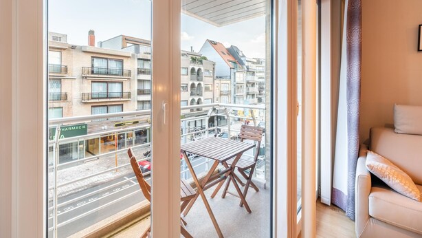 Ein Balkon mit Tisch und Stühlen, einer Couch und Blick auf eine Stadtstraße mit Gebäuden und einer Apotheke.
