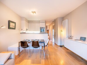 A modern kitchen with white cabinets, a central island, and a dining area with a table and chairs. - Nice apartment with balcony in the village (Koksijde)
