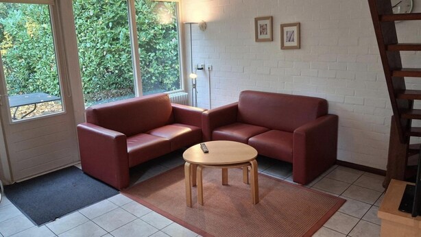 A room with a red leather sofa, a small wooden table, and a view of greenery through a window. - Bright 5P Bungalow in Limburg (Simpelveld)