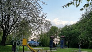 A playground with a yellow slide and climbing structures, surrounded by trees and a blue building in the background. - Bright 5P Bungalow in Limburg (Simpelveld)