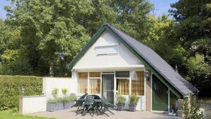 A single-story house with a sloped roof, a front porch with a table and chairs, and a small garden area with potted plants. - Bright 5P Bungalow in Limburg (Simpelveld)