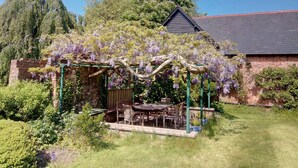 Un jardín con un pérgola cubierta de enredaderas de glicinia, un muro de ladrillo y una mesa de madera con sillas.