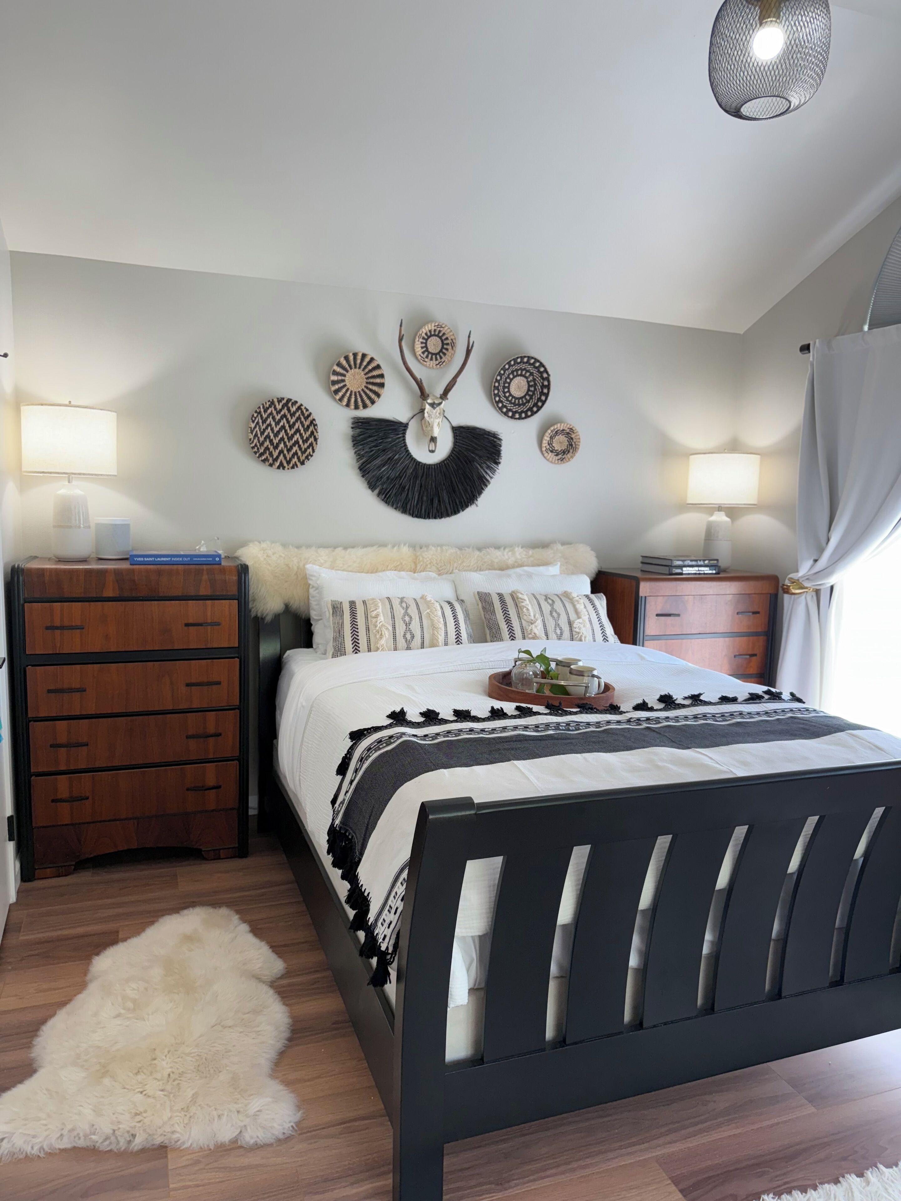 A bedroom with a bed, wooden nightstands, a fluffy rug, and wall decor including a wall-mounted animal skull and patterned wall hangings.