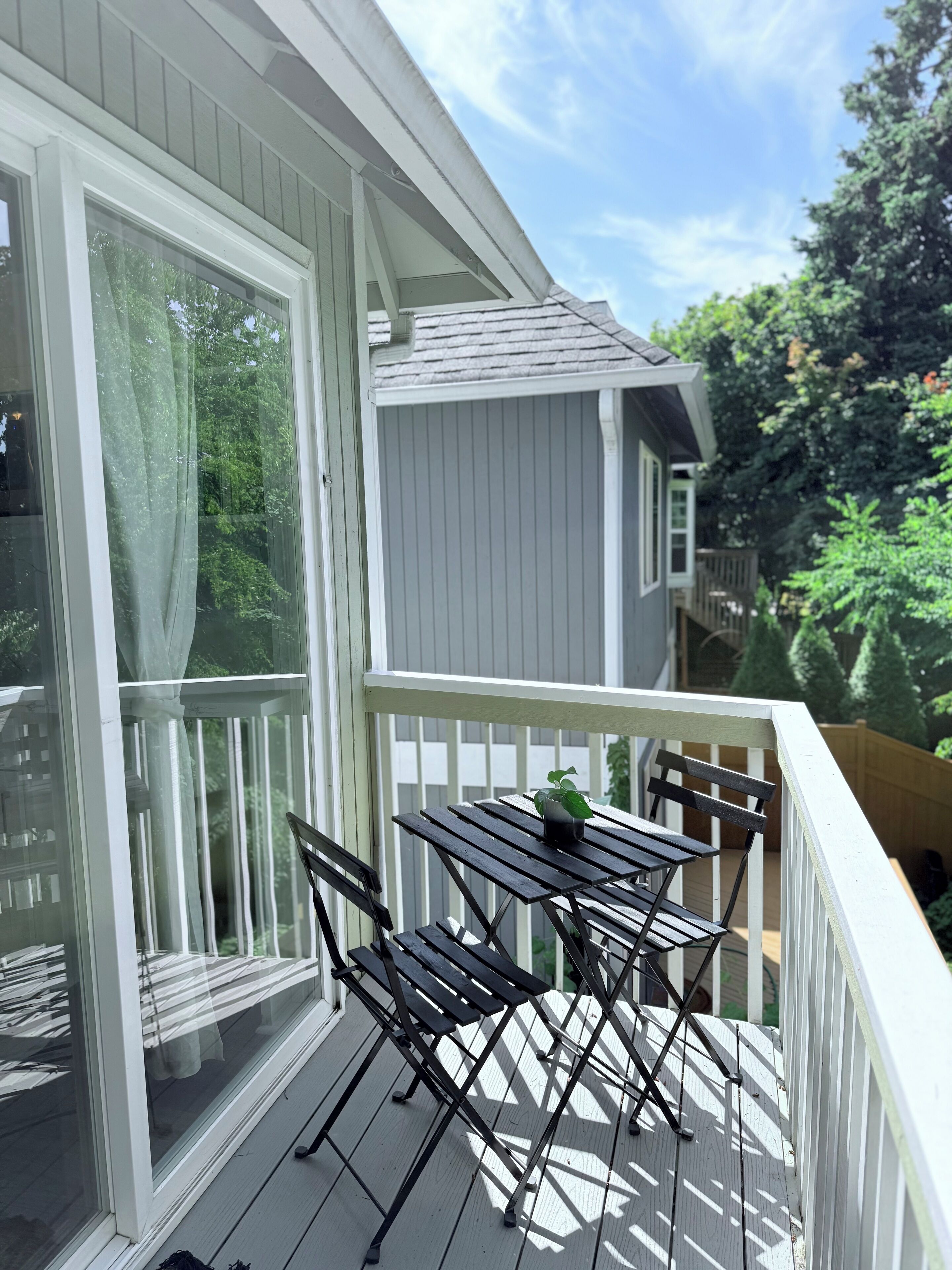 A balcony with a table and chairs, a potted plant, and a view of trees and a neighboring house.