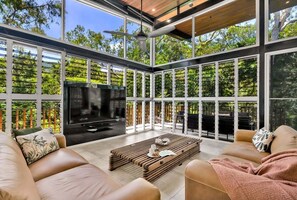 A modern living room with a large TV, comfortable seating, and large windows offering a view of greenery. - Belle Escapes Rainforest Shore (Palm Cove)