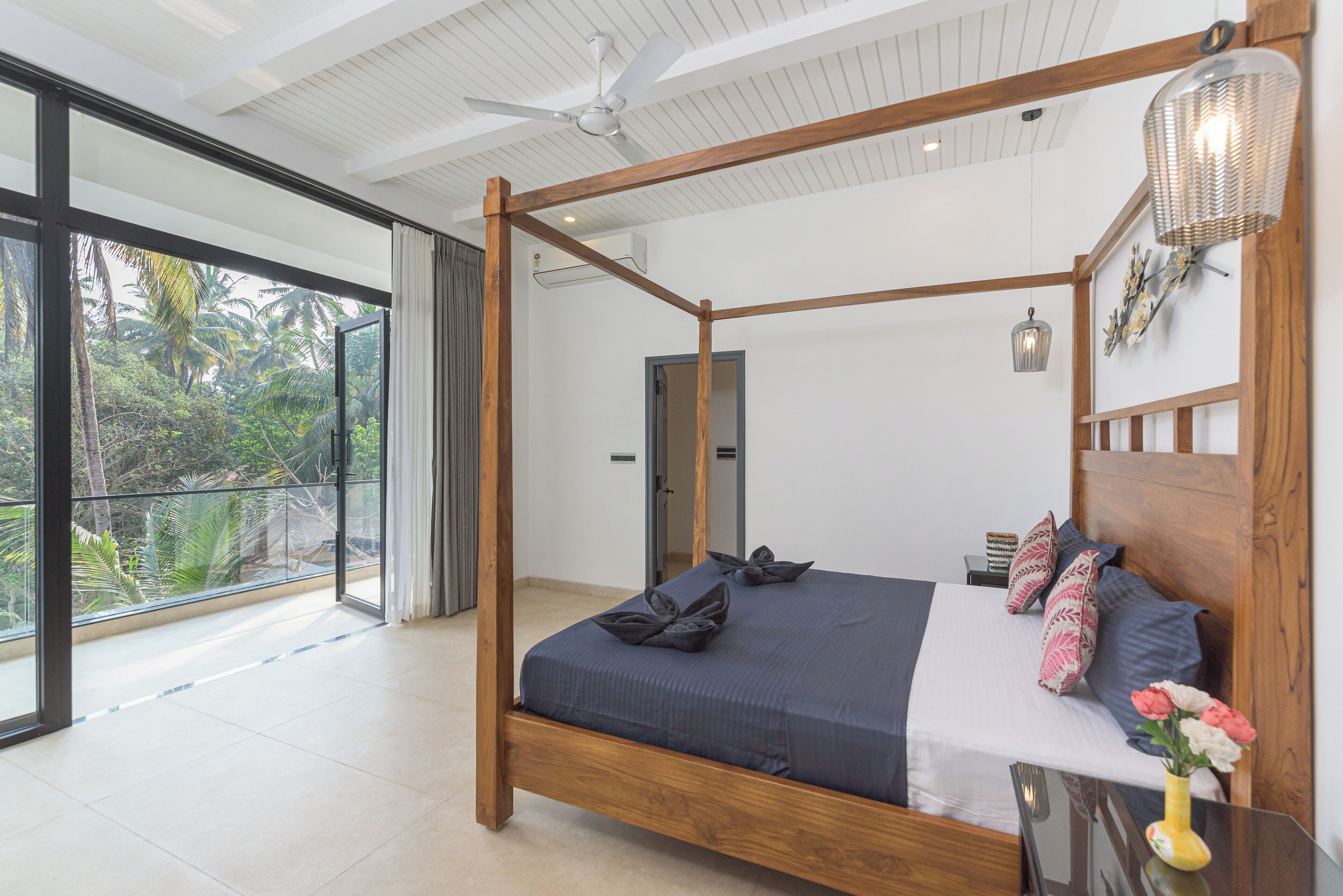 A spacious bedroom with a large bed, a balcony with a view of greenery, a ceiling fan, and a decorative wall lamp.