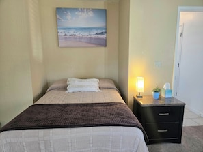 A bedroom with a bed, a bedside lamp, a nightstand, and a painting of a beach scene. - Private Guest Suite + Pool Access, North Hanford (Hanford)