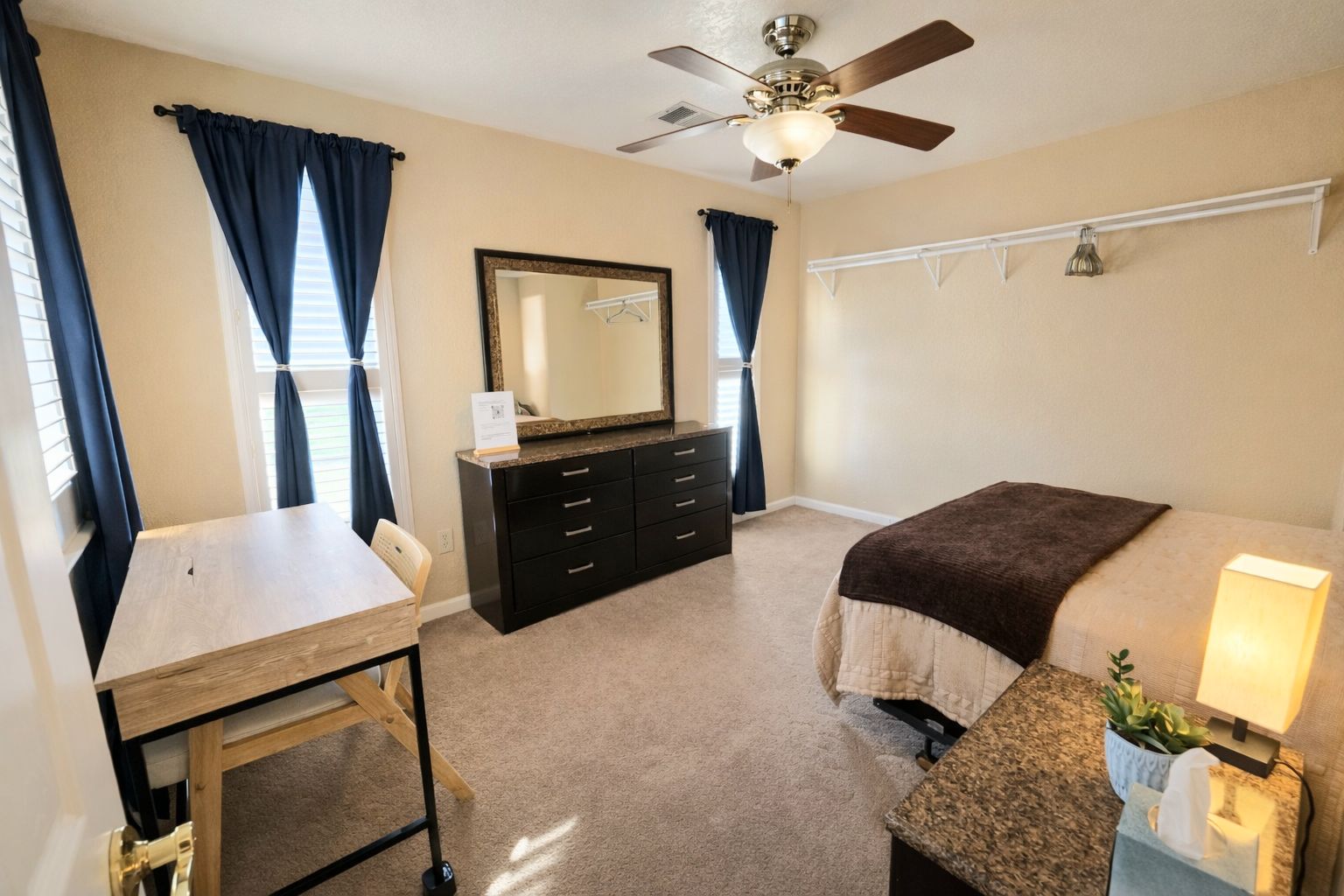 A bedroom with a bed, a desk, a dresser, a mirror, a ceiling fan, and a bedside lamp.