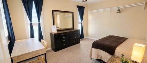 Une chambre à coucher comprenant un lit, un bureau, une commode, un miroir, un ventilateur de plafond et une lampe de chevet.