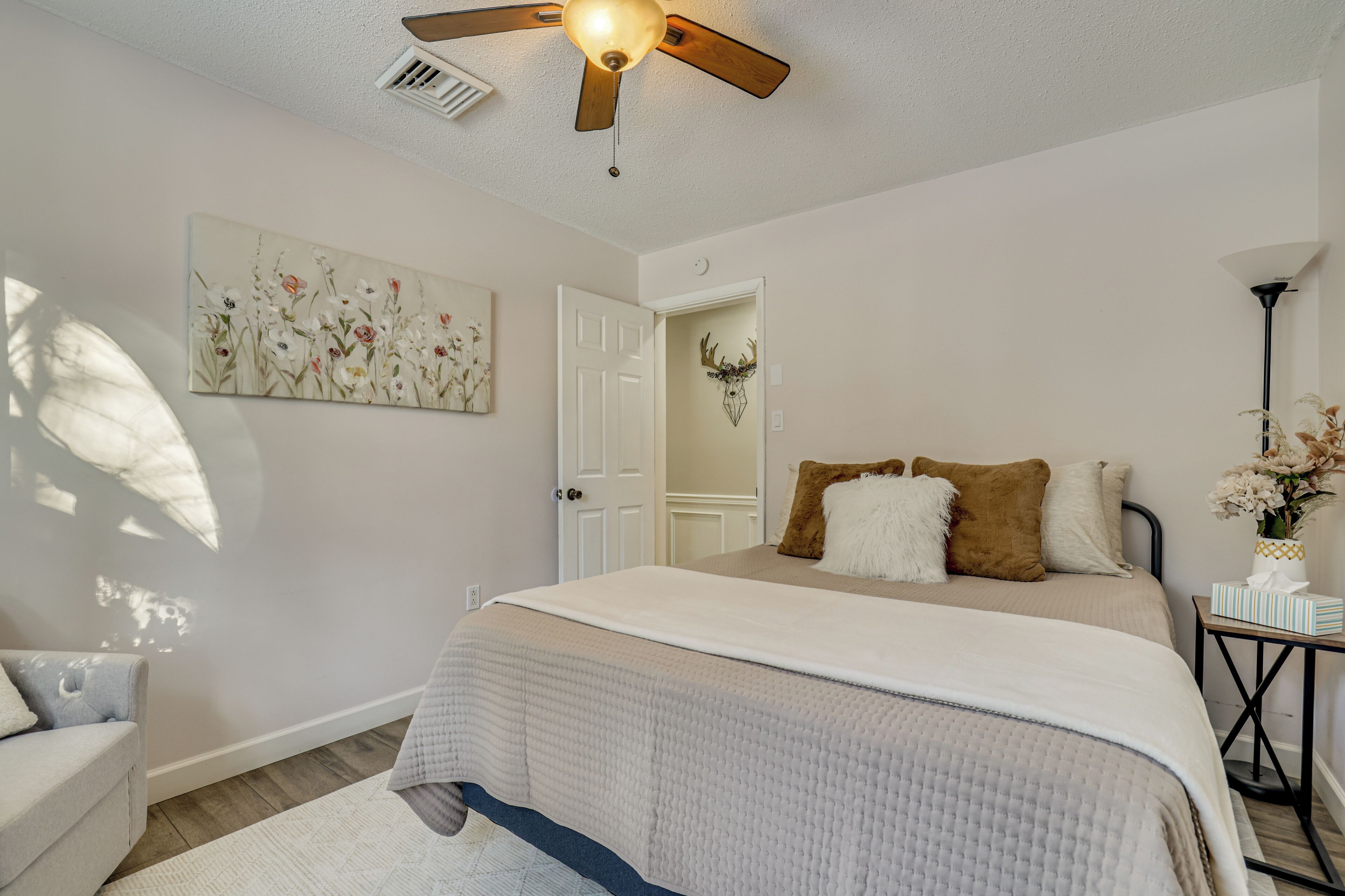 A bedroom with a bed, a ceiling fan, a door, a wall-mounted artwork, and a bedside table with a vase.