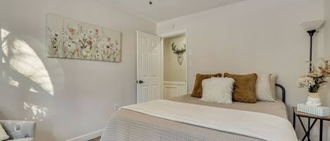 A bedroom with a bed, a ceiling fan, a door, a wall-mounted artwork, and a bedside table with a vase.