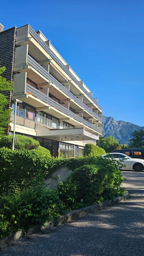 A multi-story building with balconies, a parking area, and a mountainous backdrop.