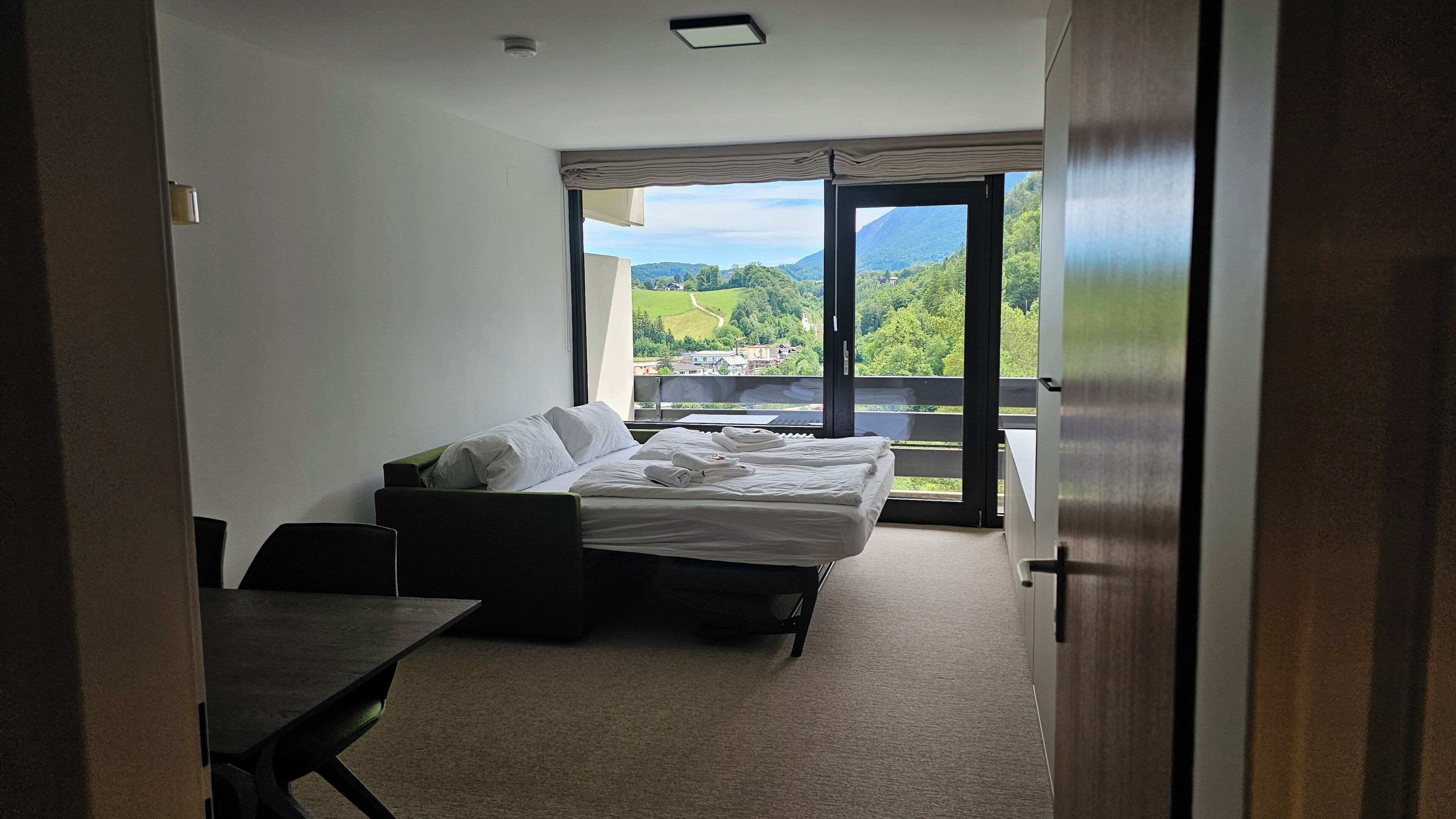 A hotel room with a bed, a small table, and a view of a green landscape through a large window.
