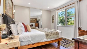 A bedroom with a large bed, a wooden nightstand, a mirror, and a window with a view of trees.