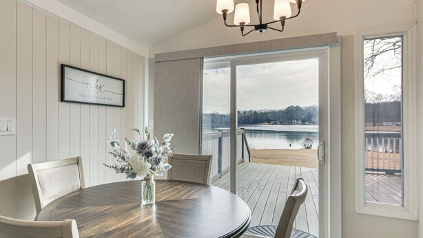Ein Essbereich mit einem runden Holztisch, Stühlen, einer Vase mit Blumen und Blick auf einen See durch große Schiebeglastüren.