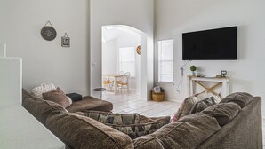 Uma sala de estar com sofá, televisão de tela plana na parede e um espaço de jantar ao fundo.