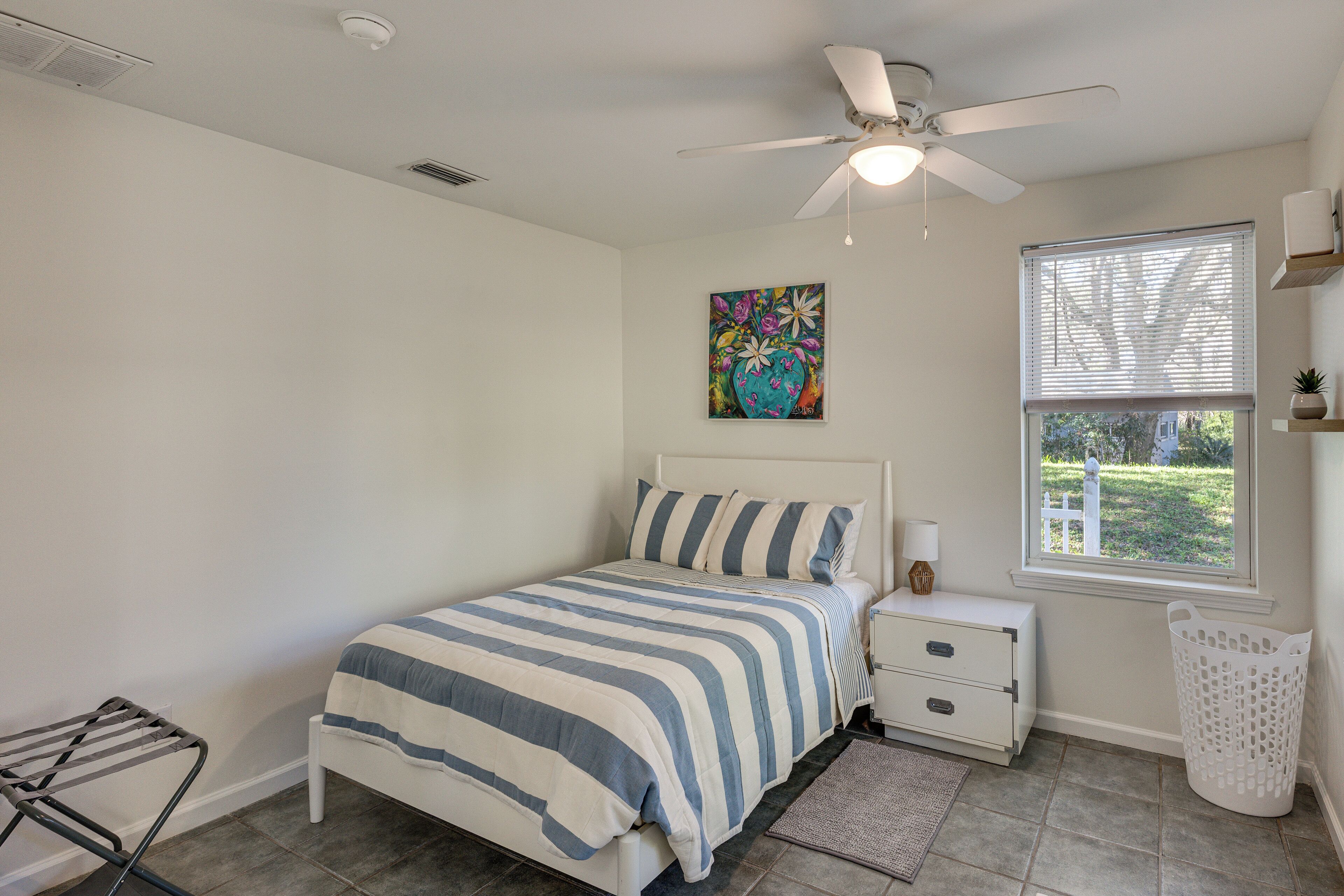 Un dormitorio con cama, mesita de noche, decoración en la pared, ventana y ventilador de techo.