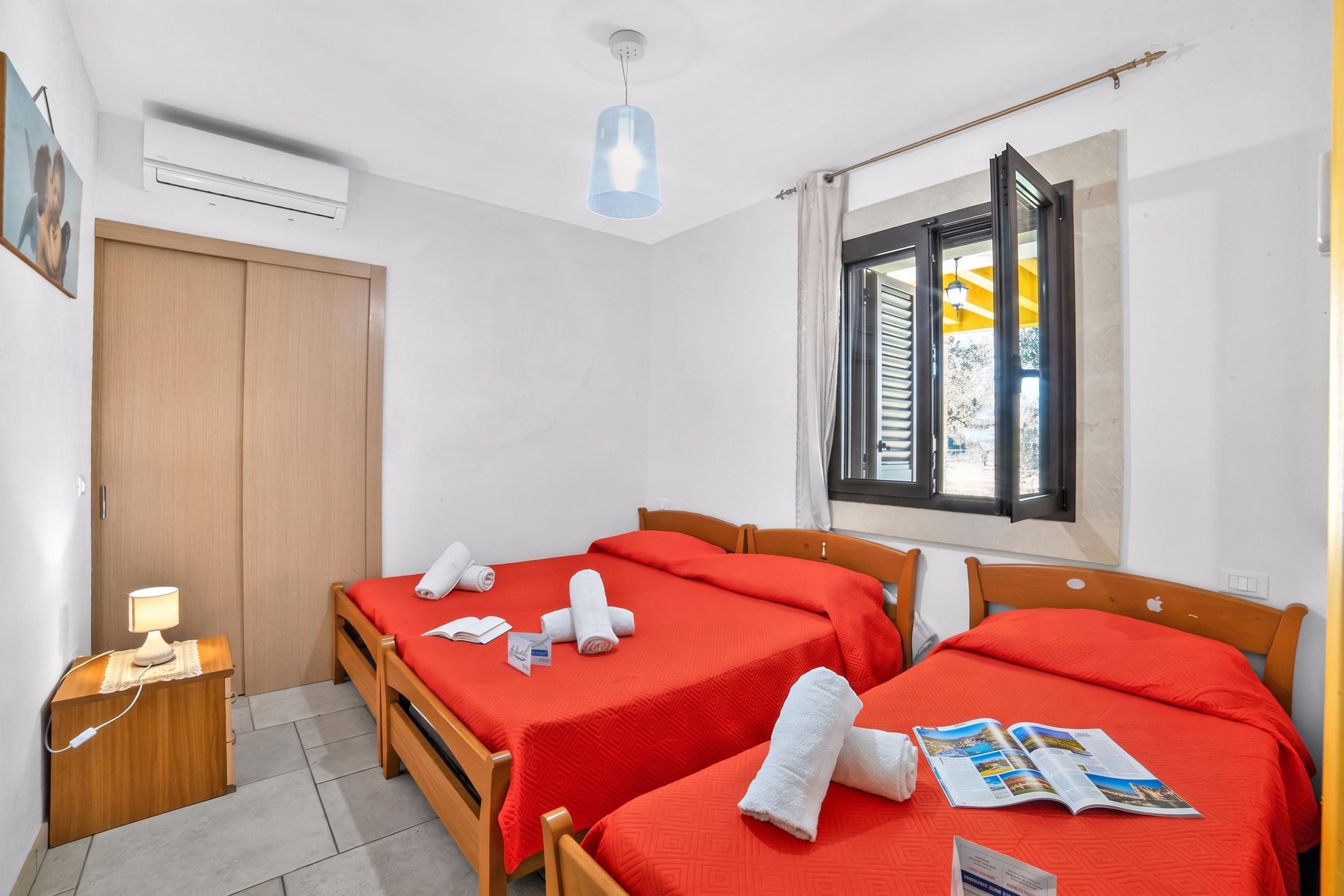 A hotel room with two beds, a window with shutters, a bedside table with a lamp, and an air conditioning unit on the wall.