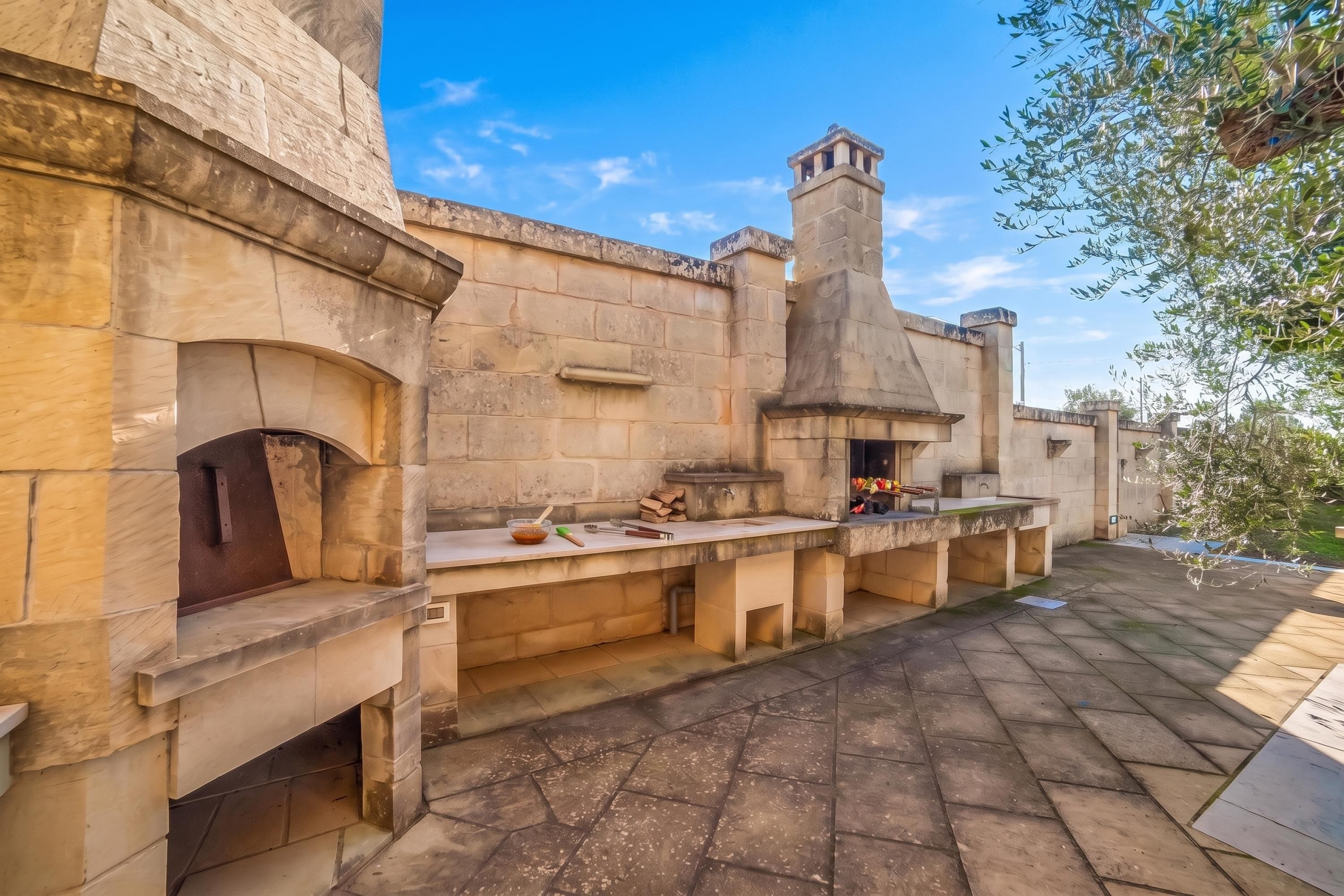 An outdoor kitchen area with stone walls and a chimney, featuring a countertop with cooking utensils and a bowl.