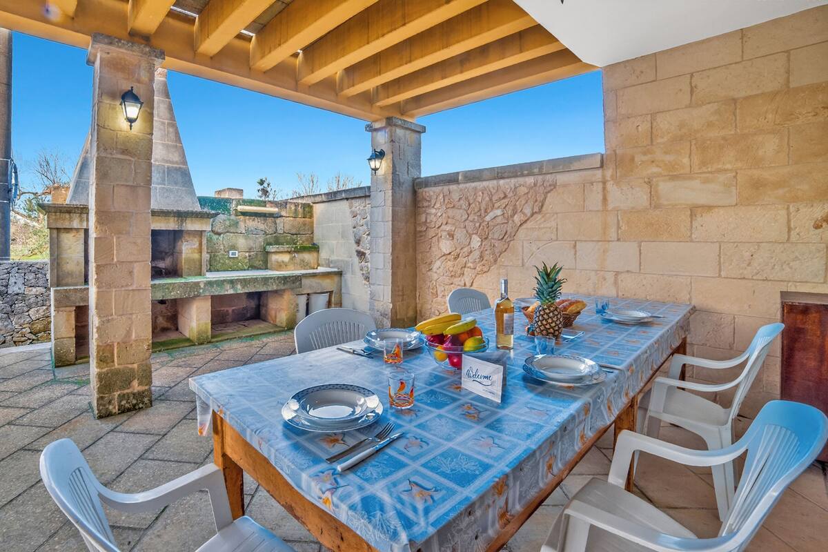 An outdoor dining area with a stone barbecue, a long table set with plates, cutlery, and a fruit bowl.