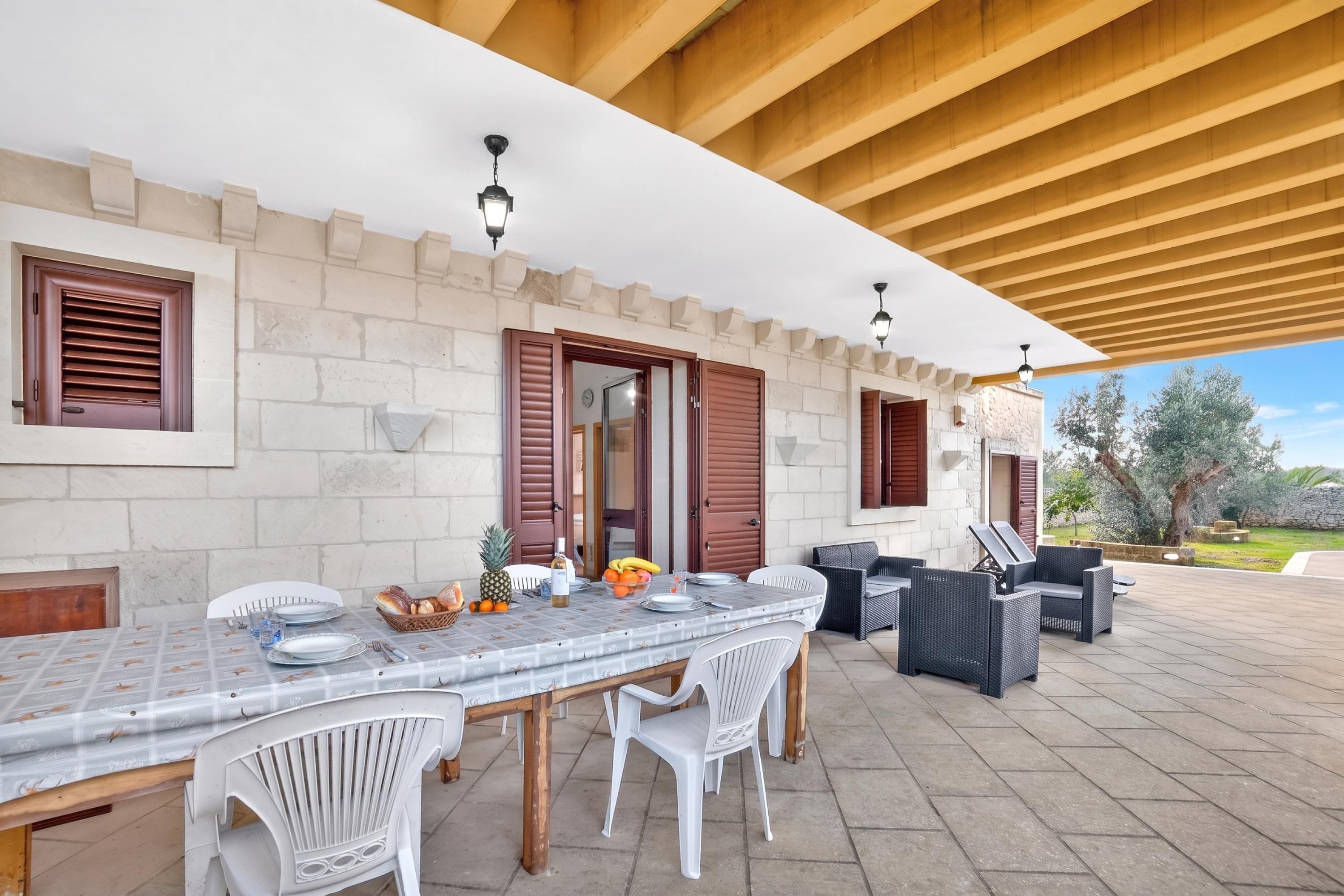 Un patio exterior con una mesa de comedor para cuatro personas, rodeado de sillas blancas, bajo una pérgola de madera.