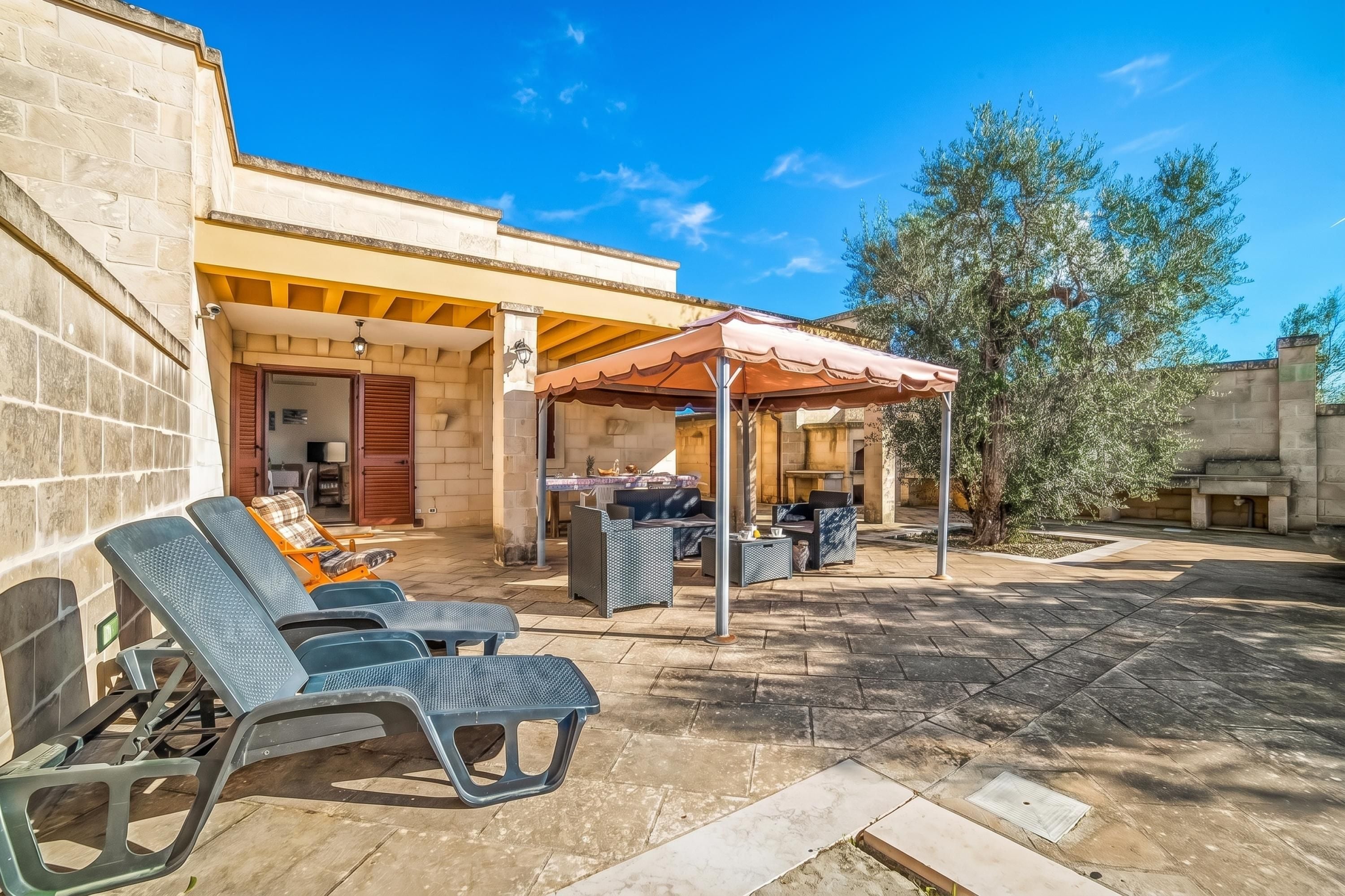 Un espace extérieur abrité avec des fauteuils et une table, un bâtiment en pierre doté d’une terrasse carrelée, et un arbre en arrière-plan.