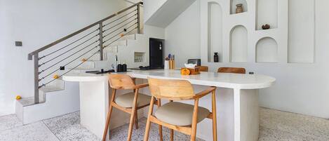 A modern kitchen with a central island, wooden chairs, a staircase, and a ceiling fan.