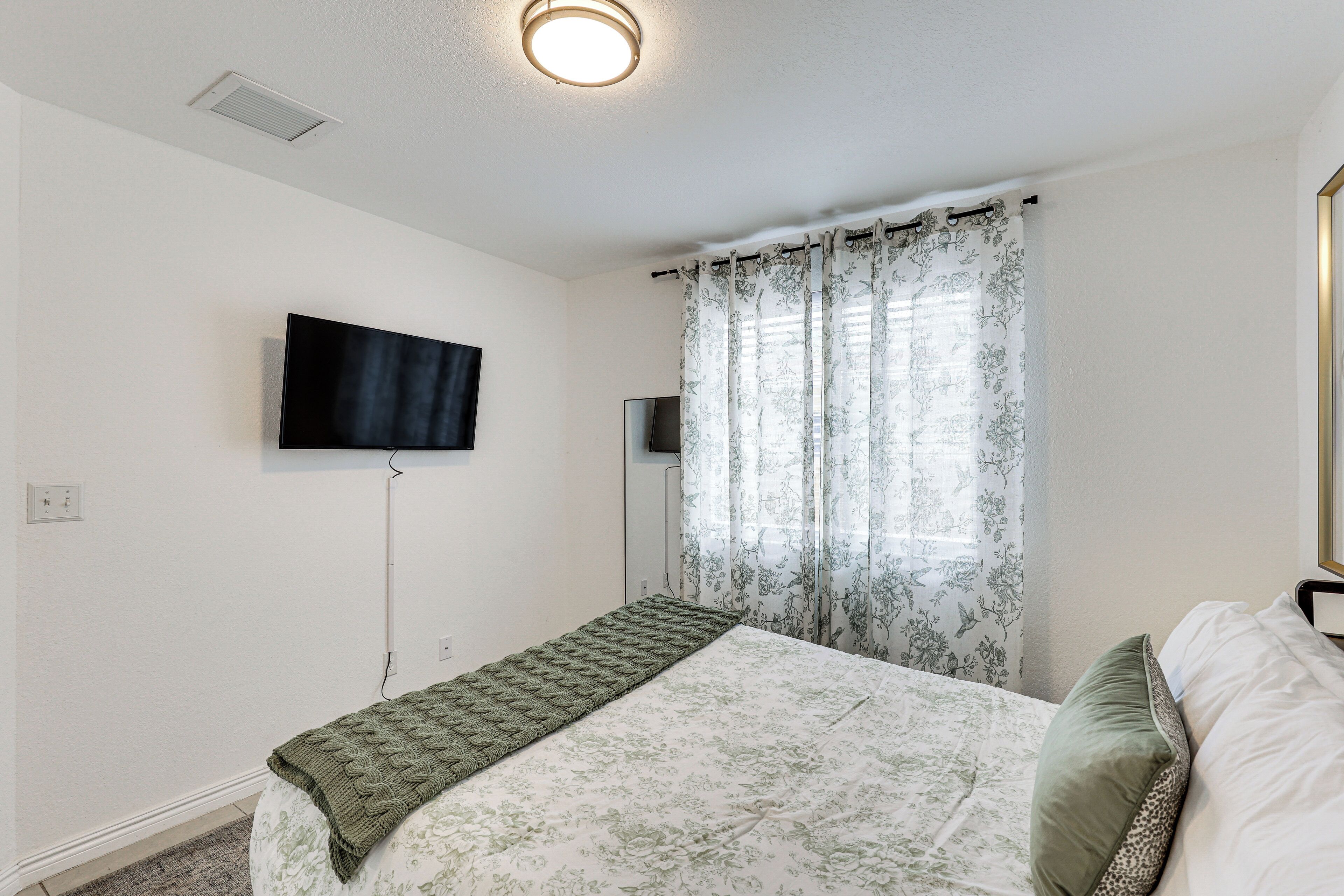 A bedroom with a bed, a television mounted on the wall, and a window with floral curtains.