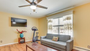 A living room with hardwood flooring, a grey sofa, a wooden coffee table, a ceiling fan, and a TV mounted on the wall. - Pet Friendly & WFH Ready! Quiet Hudson Valley Apt (Newburgh)