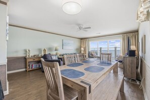 Une salle à manger spacieuse avec un grand ventilateur de plafond, une table en bois agrémentée de sets de table bleus, et une vue sur l’océan grâce à de grandes fenêtres.