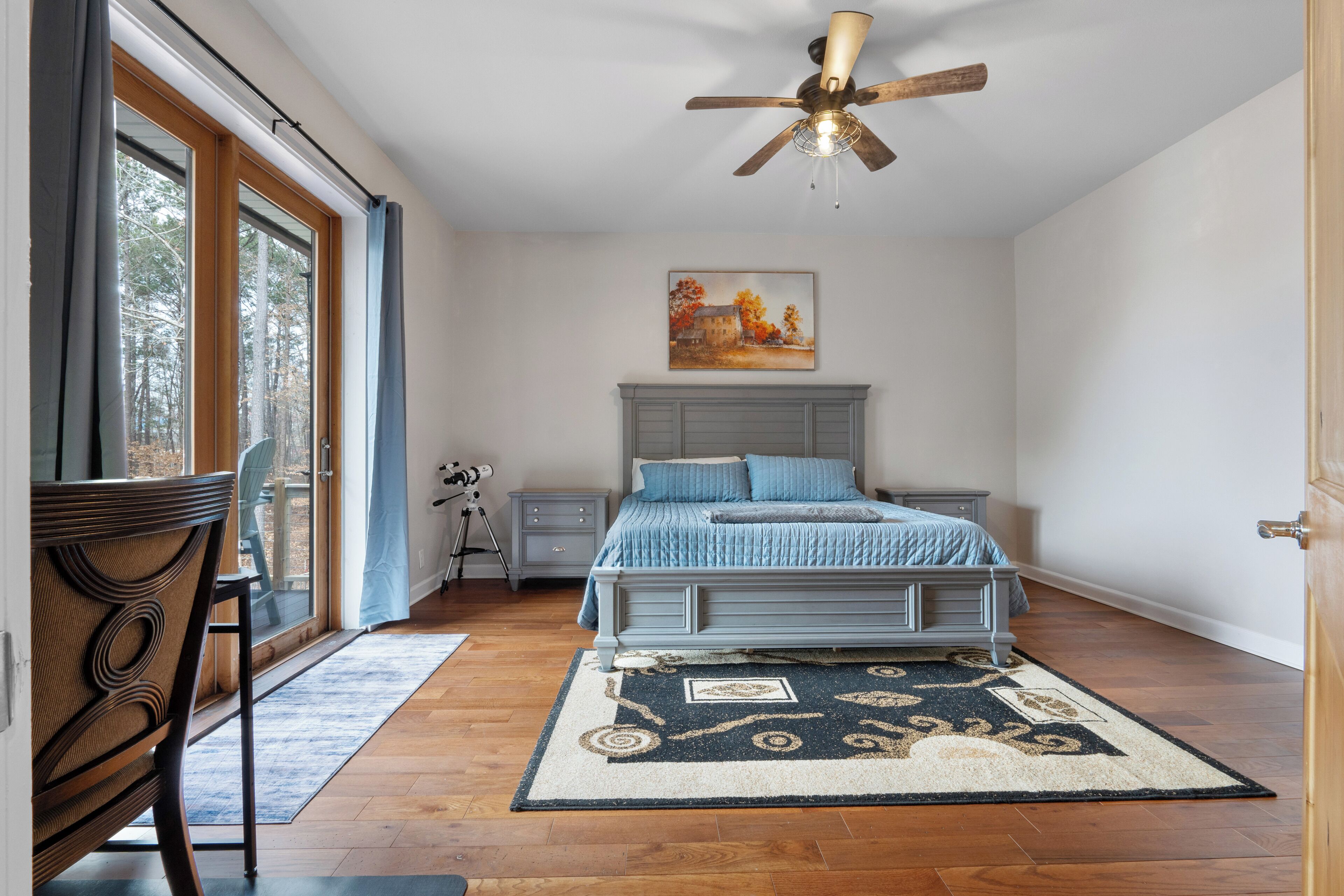 A bedroom with a bed, a ceiling fan, a nightstand, a chair, a window with curtains, and a painting on the wall.