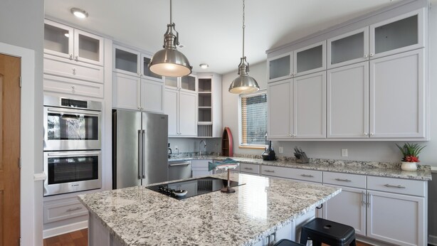 Cozinha moderna com armários brancos, eletrodomésticos de aço inoxidável, bancadas de granito e iluminação pendente.