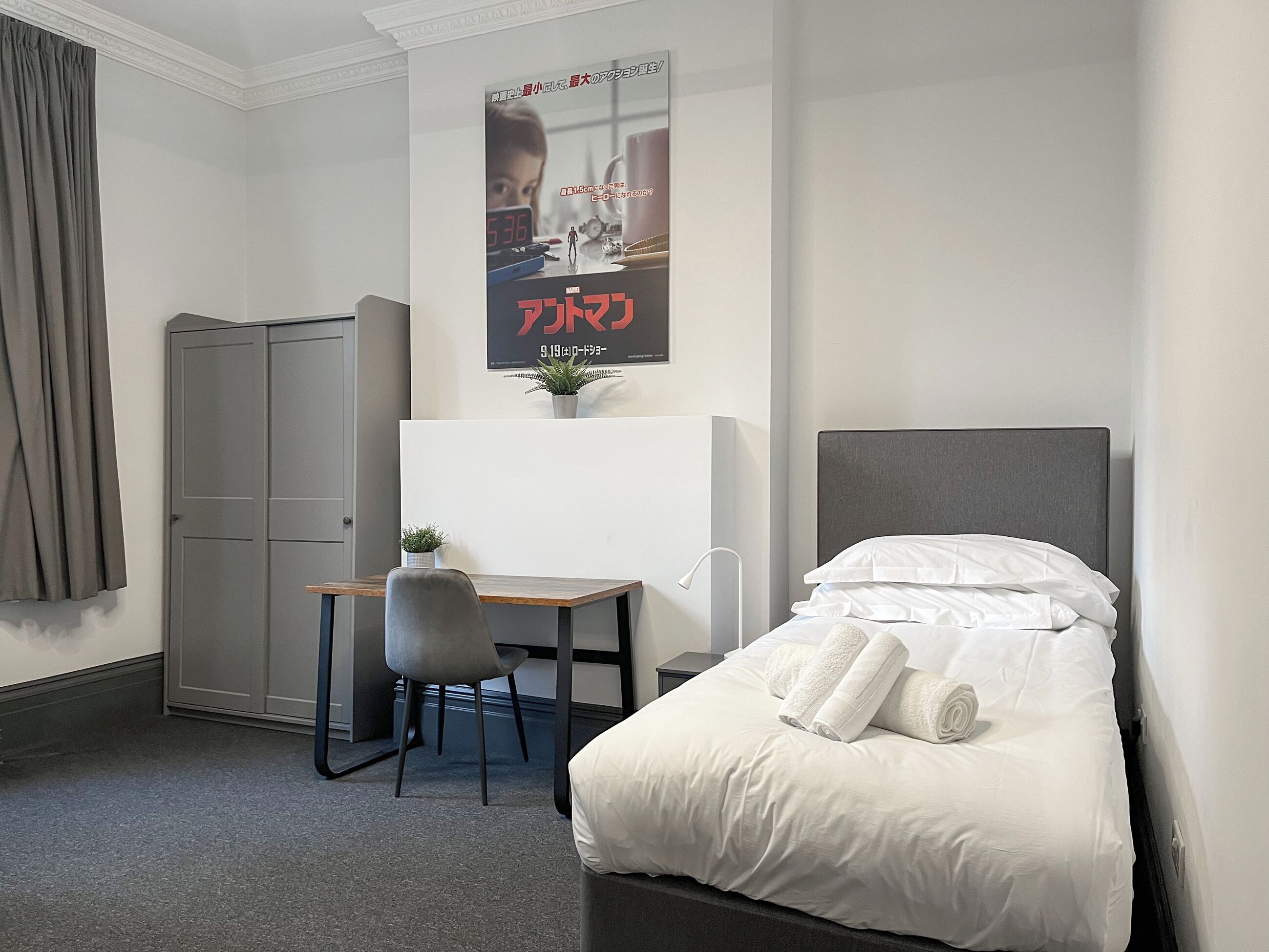 A hotel room with a bed, a desk, a chair, a wardrobe, and a poster on the wall.