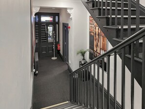 Un couloir avec un escalier, une porte dotée d’une fenêtre, un extincteur et une petite plante posée sur une table.