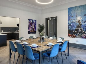 Un coin repas avec une table en bois dressée pour six personnes, des chaises bleues et une grande affiche accrochée au mur.