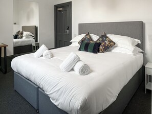 Un lit bien fait, avec des draps blancs, deux coussins décoratifs et des serviettes enroulées posées sur le lit.