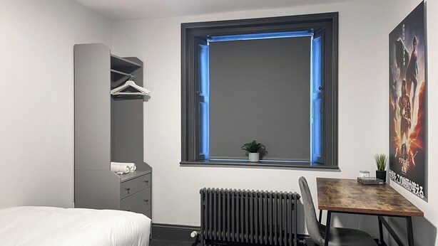 A bedroom with a bed, a desk, a chair, a radiator, a window with blue lighting, a coat rack, and a poster on the wall. - 8 bedroom accommodation in Sunderland (Sunderland)