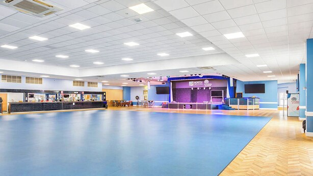 A spacious indoor area with a reception desk, a stage with a sign, and a large open floor space. - 68G Falcon's Reach - UKC8997 (Illfracombe)