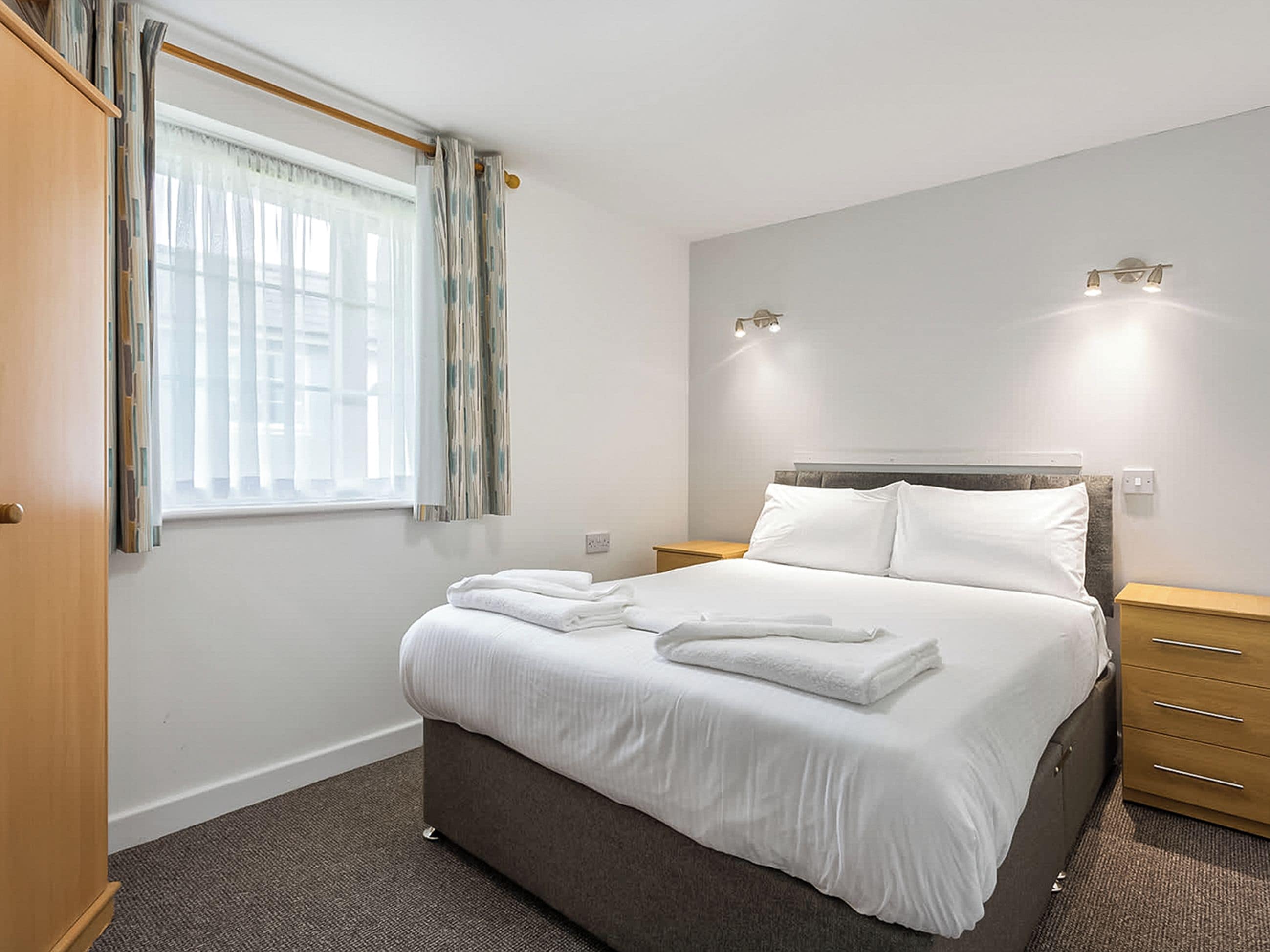 A hotel room with a bed, a window with curtains, a bedside table, and a wall-mounted light fixture.